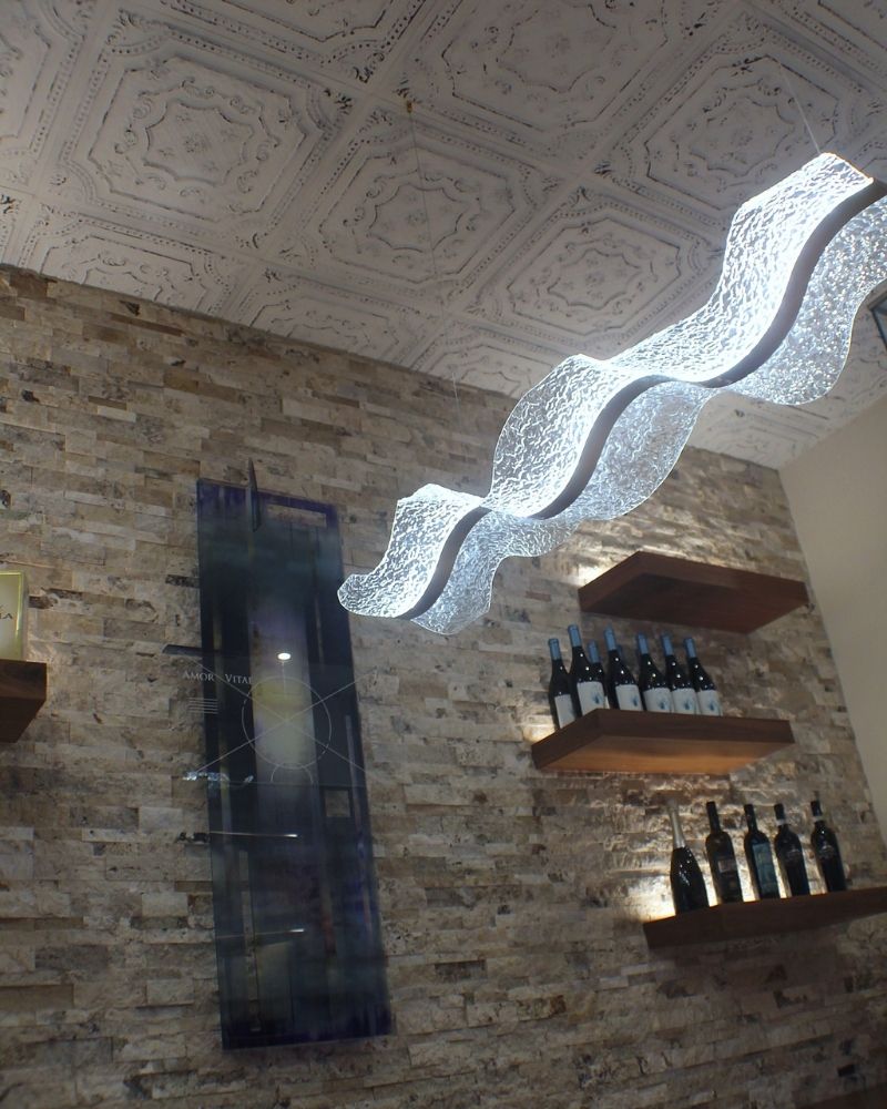 Elegant bar interior featuring a unique wavy light fixture, stone wall, and shelves displaying various wine bottles.