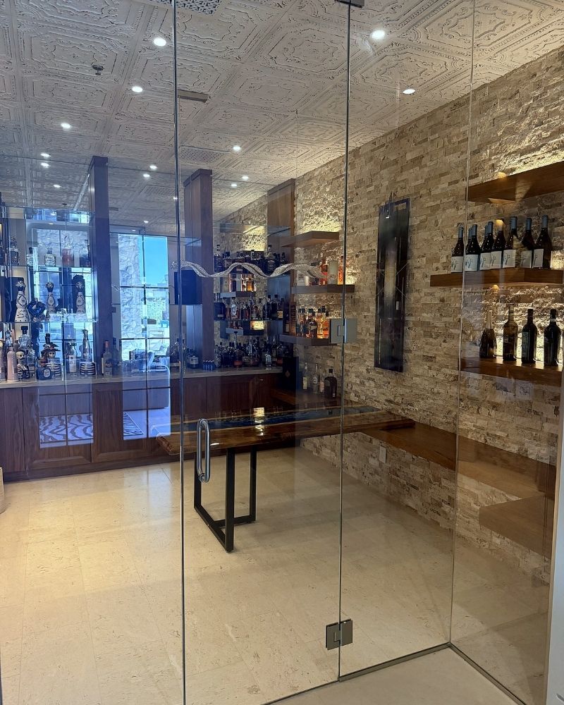 Modern bar interior with a glass entrance, showcasing an array of bottles on wooden shelves and elegant decor.
