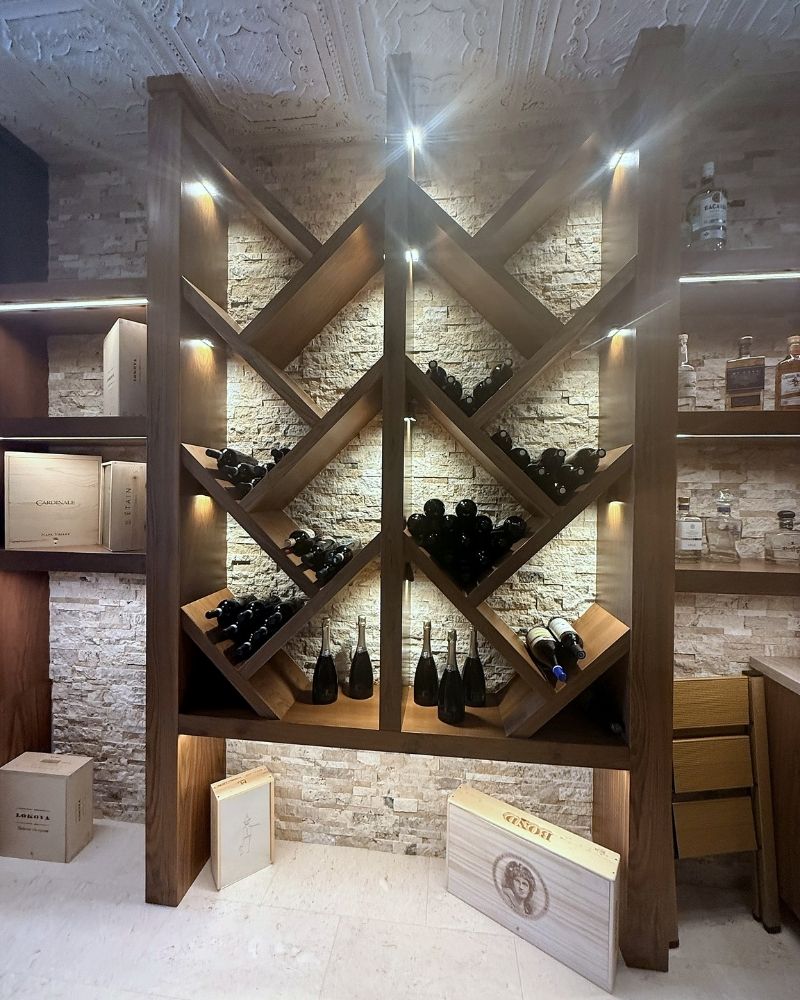 Elegant wine rack display with bottles and wooden boxes, illuminated against a stone wall in a stylish interior.