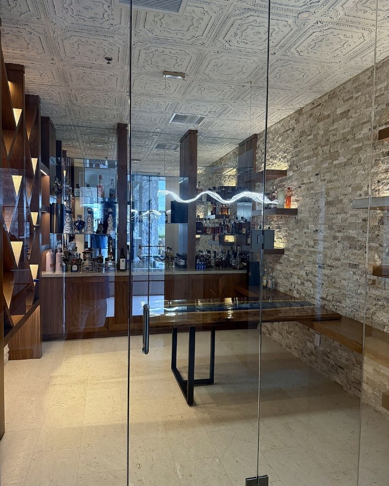 Modern bar area with glass walls, elegant shelving, and stylish decor showcasing various spirits and beverages.