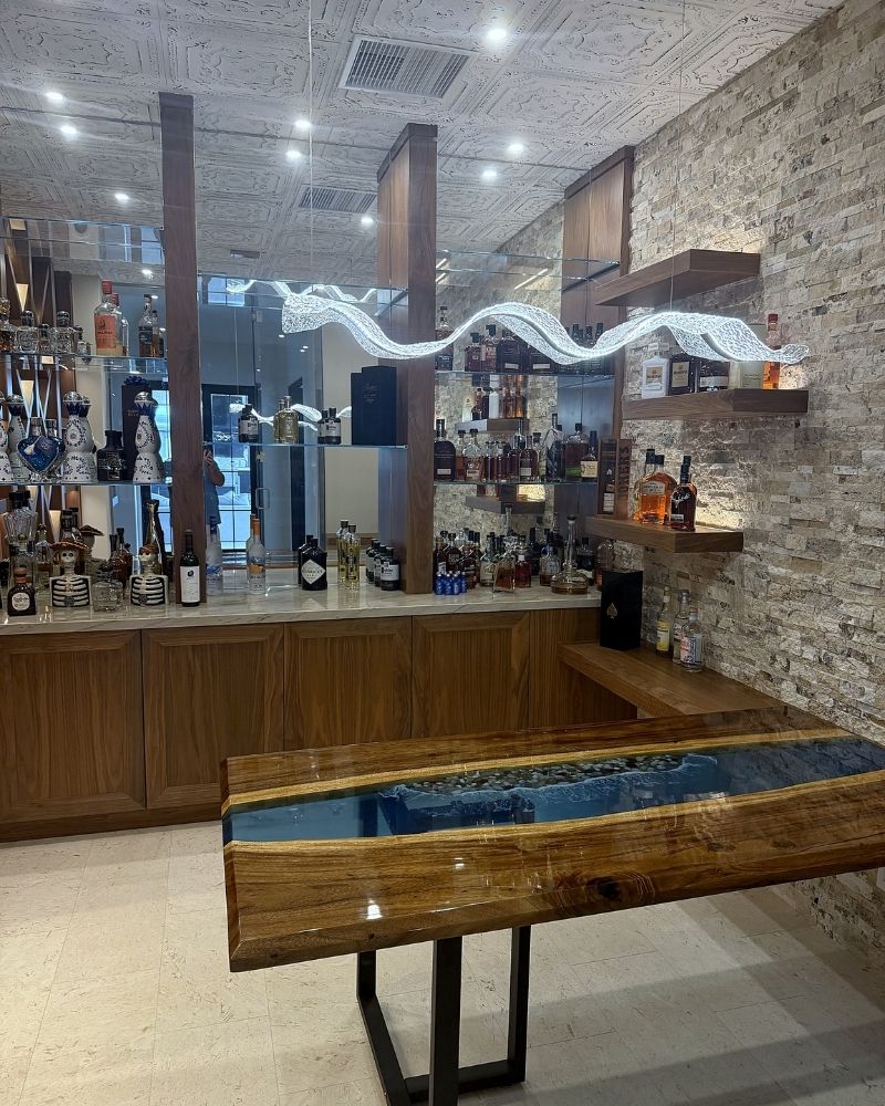 Modern bar interior featuring a wooden table with glass inlay and shelves stocked with a variety of spirits and drinks.