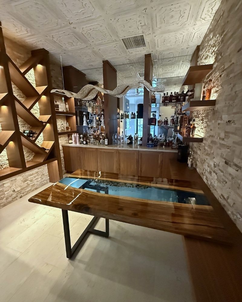 Completed Wine Cellar (38) Alt text: Modern bar interior featuring a unique wooden table with a glass section, creative lighting, and a stylish shelf display.
