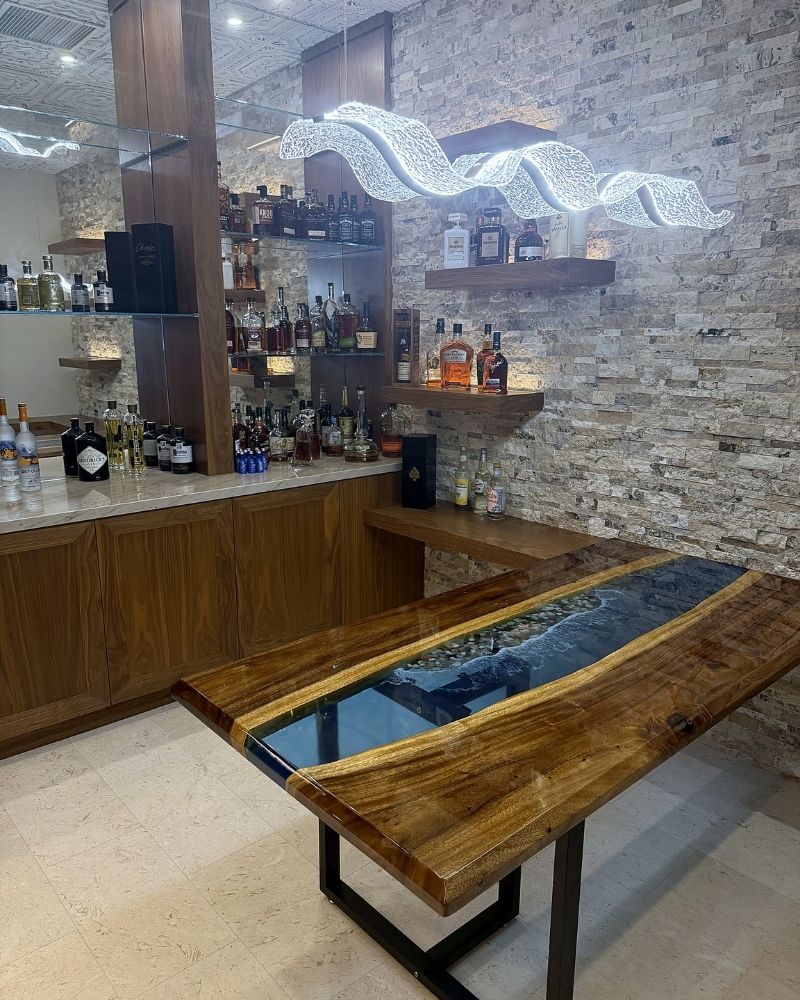 Stylish home bar featuring a stunning wooden table with a resin inlay, illuminated by a modern wave-shaped light fixture.