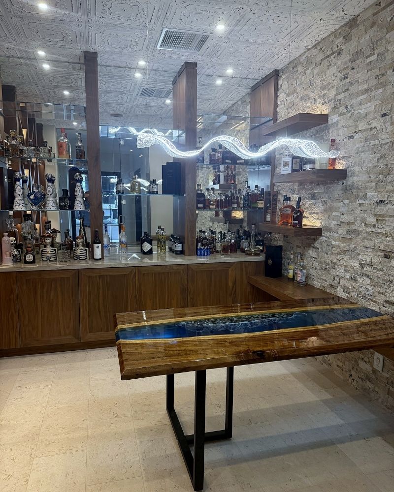 Modern home bar featuring a wood table with blue resin inlay, illuminated shelves, and a variety of liquor bottles.