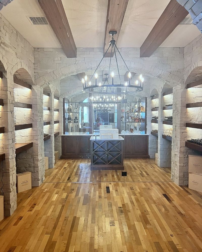 Elegant wine cellar featuring stone walls, wooden beams, and a central tasting area, illuminated by a stylish chandelier.