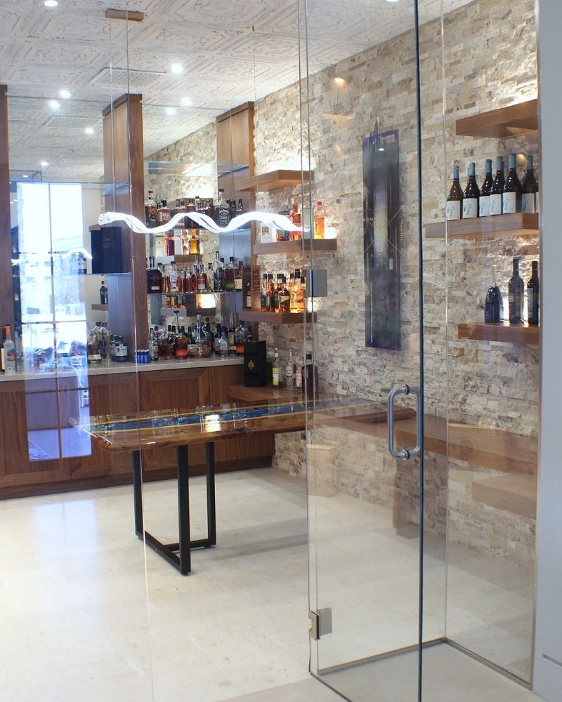 Modern home bar with a stone wall, showcase shelves, and an elegant glass entrance, featuring various spirits.