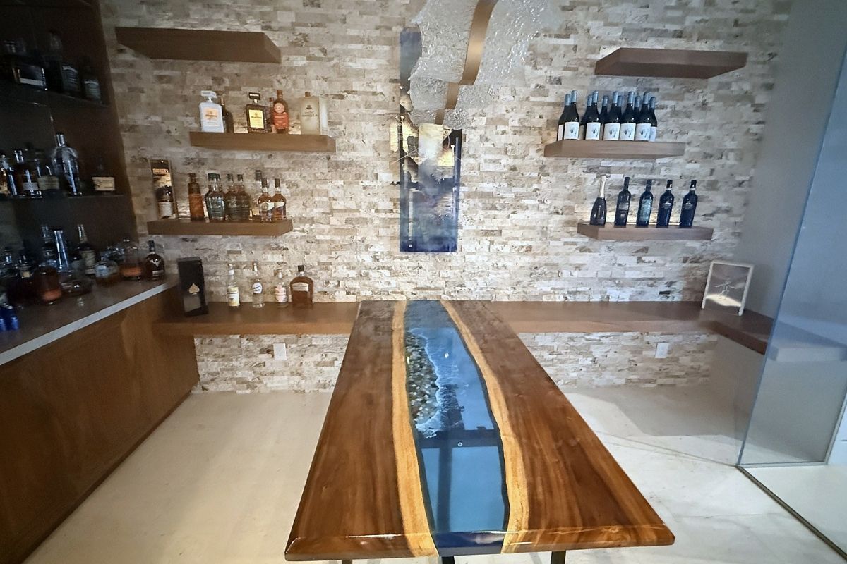 Stylish home bar featuring a wooden table with a river resin design and shelves stocked with various liquors and wine.