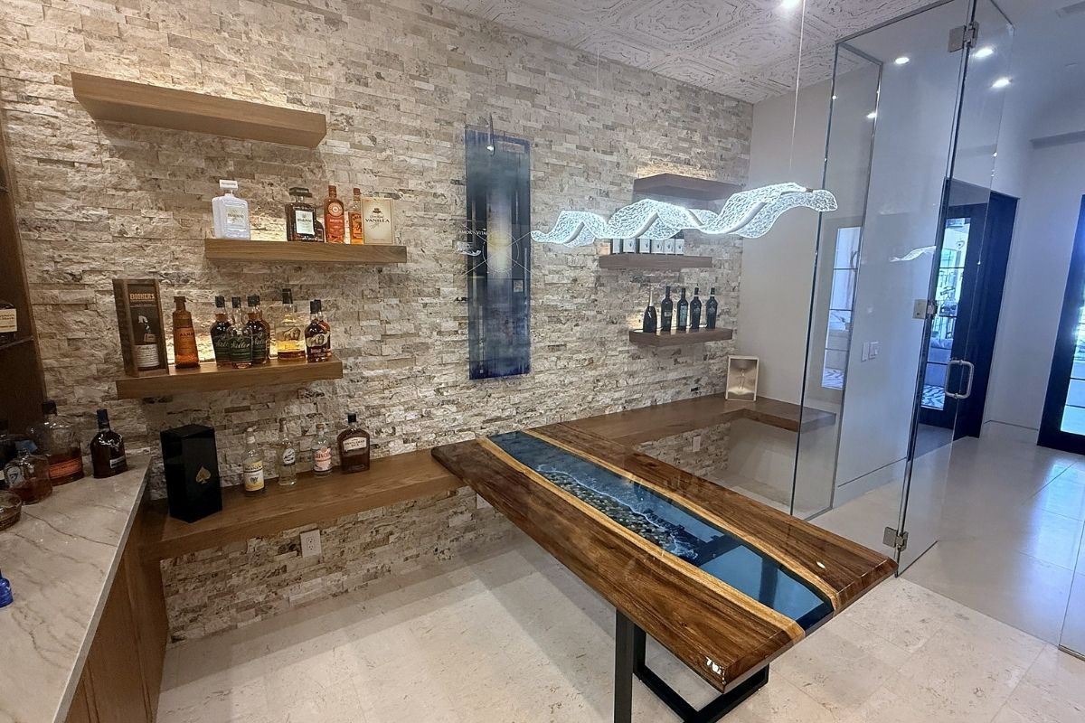 Stylish home bar featuring stone walls, wooden shelves, and a unique glass river table design.