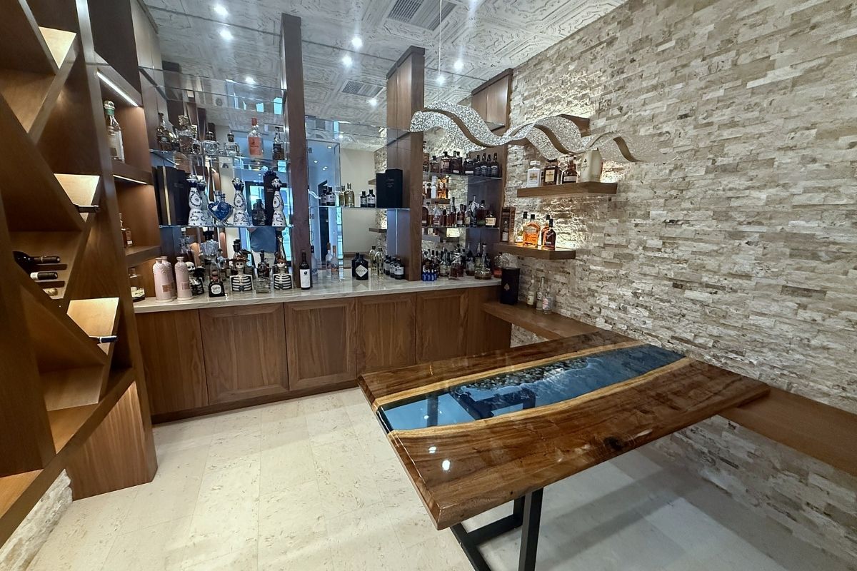 Modern bar interior featuring a polished wooden table, stacked liquor bottles, and stylish stone wall decor.