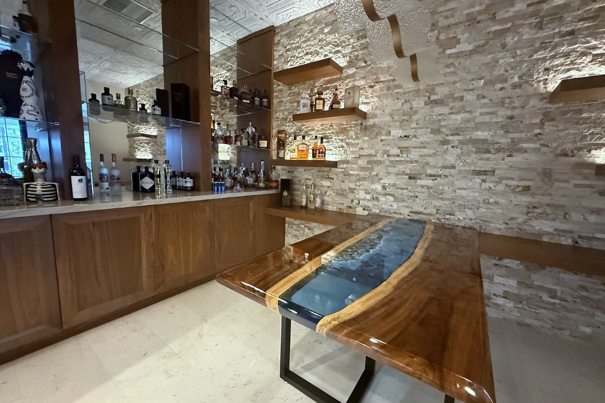 Stylish home bar with a wooden table and stone walls, showcasing an extensive liquor collection on sleek shelves.