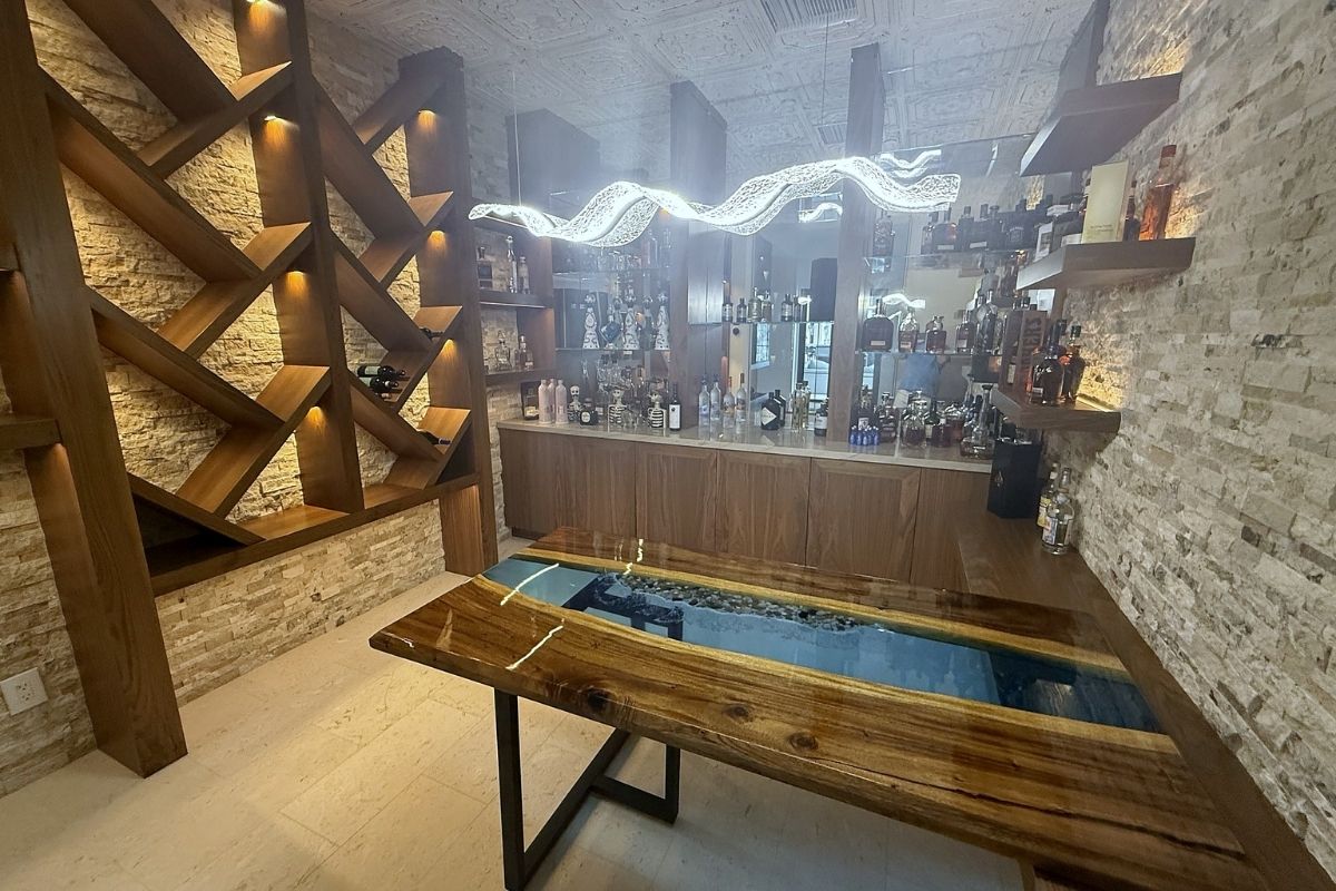Stylish home bar with illuminated shelves, a wooden table featuring a blue resin inlay, and a variety of liquor bottles.