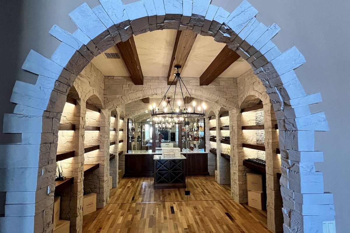 Spacious wine cellar with stone walls, wooden beams, and soft lighting, featuring shelves for bottles and a central display counter.