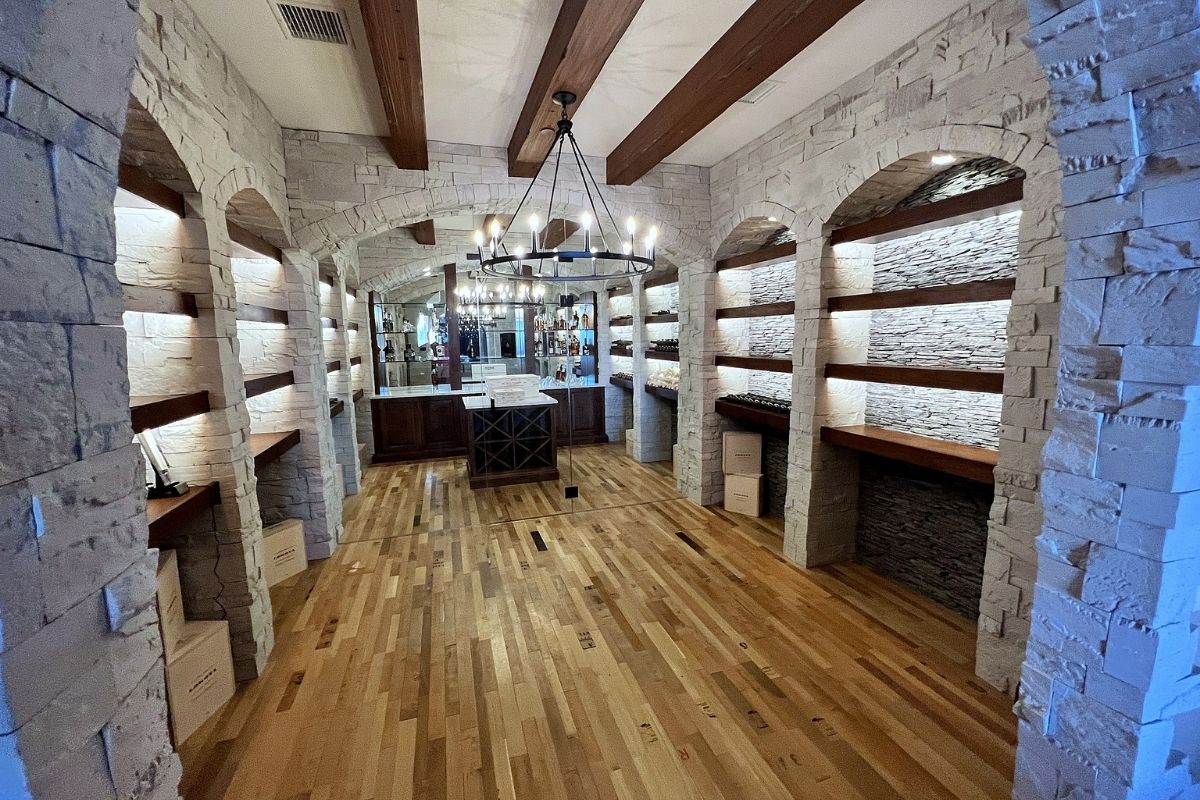 Spacious rustic wine cellar with stone walls, wooden shelves, and a modern chandelier illuminating the warm wooden floor.
