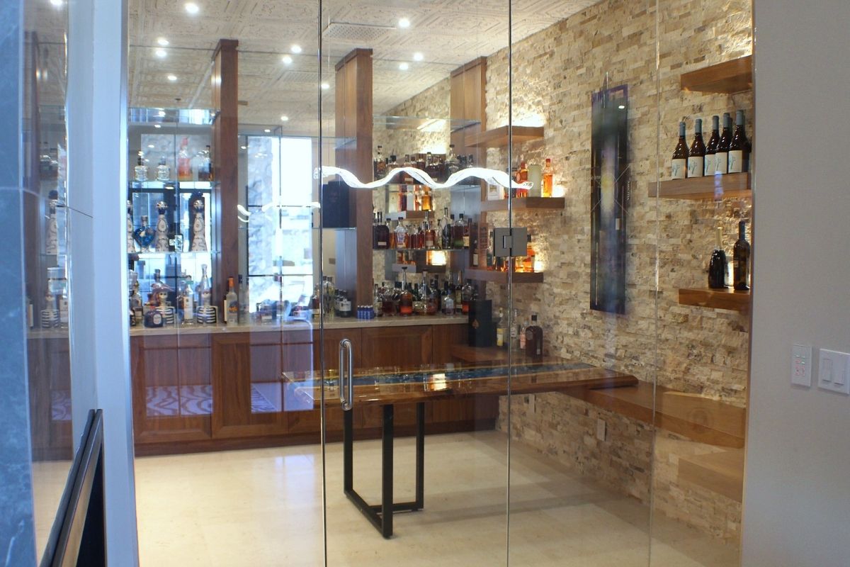 Modern home bar featuring a glass enclosure, wood shelves, and various bottles of liquor against a stone wall backdrop.