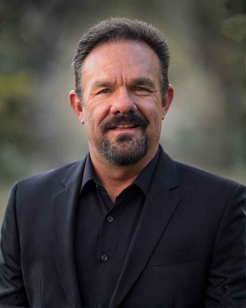 Smiling man in a black suit stands outdoors, with a blurred natural background, exuding confidence and professionalism.